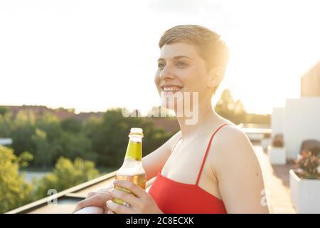 Gros plan d'une femme souriante tenant une bouteille de bière sur le balcon ciel dégagé au coucher du soleil Banque D'Images