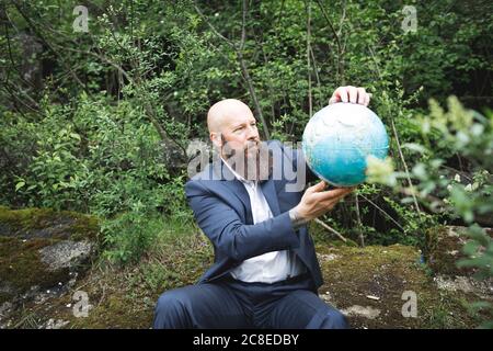 Homme d'affaires à barbe chauve tenant le globe tout en étant assis contre les arbres à l'intérieur forêt Banque D'Images