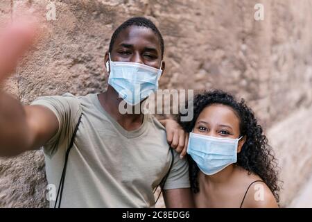 Jeune couple portant des masques tout en prenant le selfie contre le mur Banque D'Images