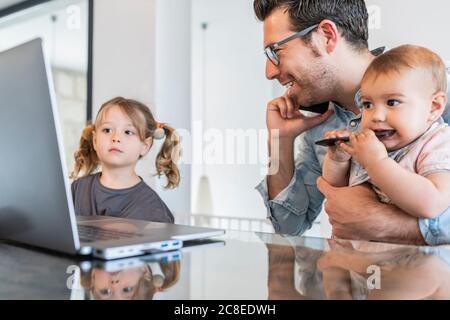 Homme tenant la fille parlant sur un smartphone pendant que la fille regarde à l'ordinateur portable sur la table Banque D'Images