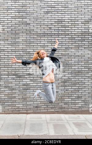Une femme heureuse avec des bras débordés sautant sur le sentier contre le gris mur de briques Banque D'Images