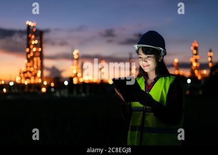 Femme asiatique ingénieur pétrochimique travaillant la nuit avec une tablette numérique à l'intérieur de l'usine de raffinage de pétrole et de gaz la nuit pour la sécurité des inspecteurs Banque D'Images