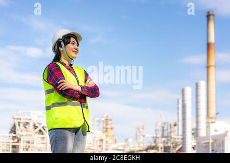 Une femme asiatique a croisé un bras et sourire avec confiance en vue de l'avenir avec l'usine de raffinerie de pétrole en arrière-plan. Banque D'Images