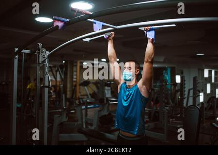 Musclé jeune homme de fitness avec masque médical faisant des exercices sur le bar horizontal dans un club de gym. Protection contre le coronavirus COVID 19 Banque D'Images