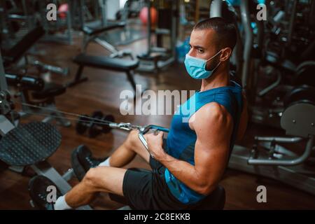 Jeune homme de forme musculaire avec masque médical faisant un exercice lourd de poids pour le dos et le bras sur la machine dans la salle de gym.prévention de COVID - 19 coronavirus Banque D'Images