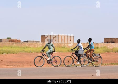 Trois adolescents à vélo sur la route principale traversant un village africain. Banque D'Images