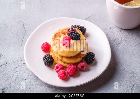 Crêpes au fromage cottage, beignets de caillé dessert aux framboises et aux mûres dans un plat près d'une tasse de thé chaud avec une tranche de citron Banque D'Images