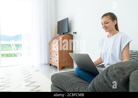 Souriante belle jeune femme assise sur un canapé en utilisant un ordinateur portable communiquant en ligne travaillant à la maison, heureuse adolescente fille dactylographiant sur l'ordinateur Banque D'Images