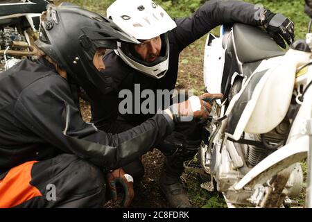 Motocyclistes concentrés dans les casques examinant le blindage inférieur de la moto tout en discutant de la raison de la panne Banque D'Images