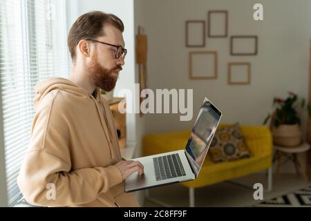 Jeune barbu concentré dans des lunettes debout à la fenêtre et analyse de l'interface croquis sur ordinateur portable Banque D'Images