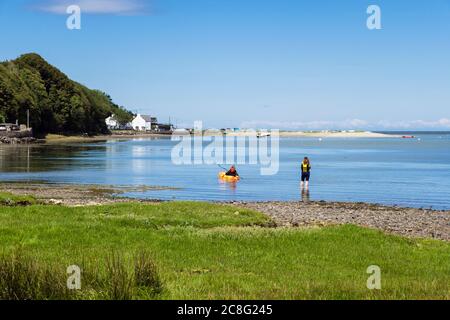 Vue sur le village avec une jeune femme pagayant un kayak dans la mer bleue calme à marée haute dans la magnifique baie de Red Wharf, île d'Anglesey, pays de Galles, Royaume-Uni, Grande-Bretagne Banque D'Images