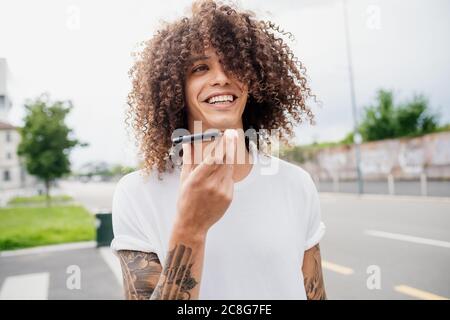 Portrait d'un homme avec des bras tatoués et de longs cheveux bruns bouclés, à l'aide d'un téléphone portable. Banque D'Images