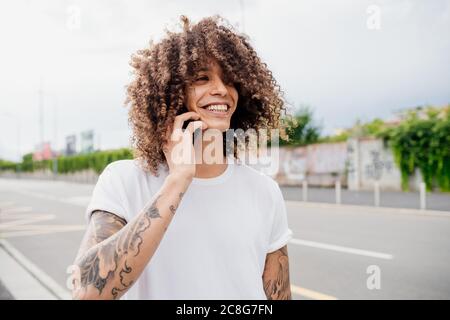 Portrait d'un homme avec des bras tatoués et de longs cheveux bruns bouclés, à l'aide d'un téléphone portable. Banque D'Images