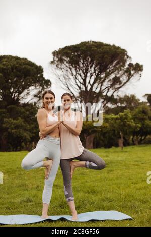 Deux femmes de fitness pratiquant le yoga dans un parc debout sur une jambe se tenant l'une l'autre. Portrait de deux femmes de forme physique debout dans l'arbre posent ensemble. Banque D'Images