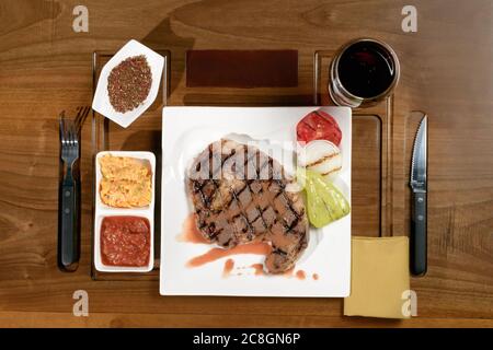 Steaks de bœuf en marbre grillé avec légumes cuits au four et épices et verre de vin rouge sur fond de bois, vue du dessus Banque D'Images