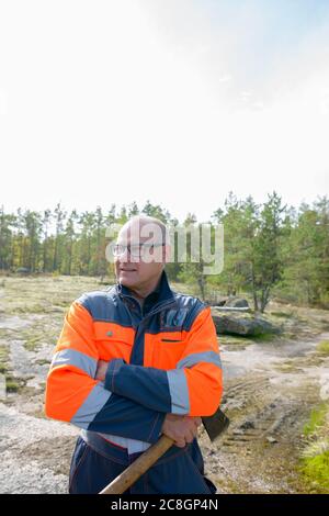 Portrait d'un homme beau et mûr heureux pensant et tenant hache dans la forêt Banque D'Images