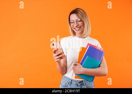 Portrait de jeune femme blonde charmante et gaie portant des vêtements décontractés, isolés sur fond orange, tenant des dossiers, à l'aide d'un téléphone portable Banque D'Images