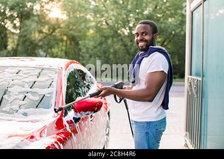 Lavage de voiture moderne rouge sur l'air libre service de lavage de voiture avec mousse et tuyau haute puissance. Jeune homme africain souriant nettoyant sa voiture avec un jet Banque D'Images