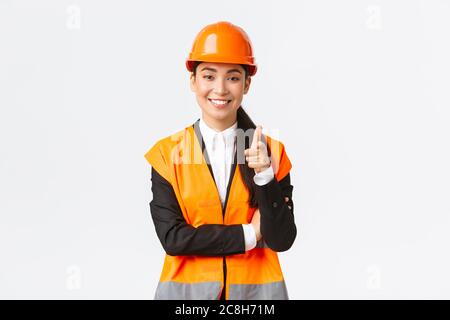 Une femme asiatique souriante et réussie, qui pointe le doigt sur la caméra, porte un casque de sécurité et une veste réfléchissante, présente la construction Banque D'Images