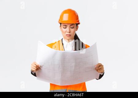 Femme asiatique confuse et douteuse ingénieur en chef, architecte principal dans la zone de construction regardant mécontente et hésitant à des plans étranges, la lecture Banque D'Images