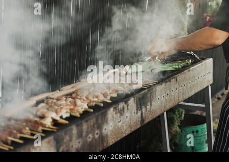 Main de jeune homme de griller de la viande et des légumes sur un long barbecue rectangulaire avec de la fumée sur la rue à l'extérieur. Barbecue d'été. Barbecue avec Banque D'Images