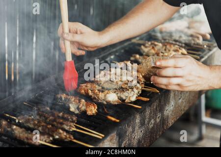 Main du jeune homme lubrifie la viande avec une brosse de sauce sur le long barbecue rectangulaire avec de la fumée sur la rue à l'extérieur. Barbecue d'été. Barbe Banque D'Images