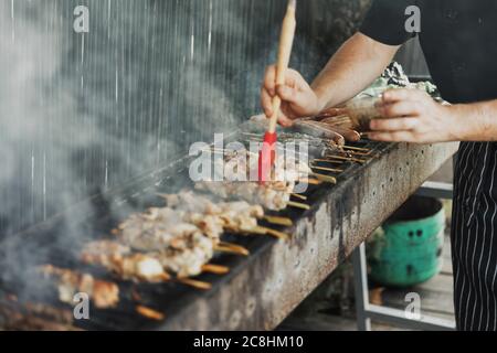 Mains floues de l'homme lubrifie la viande avec une brosse de sauce sur le long barbecue rectangulaire avec de la fumée sur la rue à l'extérieur. Barbecue d'été. BA Banque D'Images