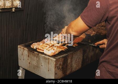 Main de jeune homme griller de la viande et du poisson sur un long barbecue rectangulaire avec de la fumée sur la rue à l'extérieur. Barbecue d'été. Barbecue avec ra Banque D'Images