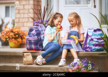 Élèves de l'école primaire avec des boîtes à lunch entre les mains. Les filles avec des sacs à dos mangent des fruits près du bâtiment à l'extérieur. Début des leçons. Premier jour de f Banque D'Images