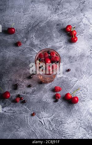 Boisson fraîche et froide à l'eau pétillante avec baies de cerisier, framboise et cassis en verre à facettes rouge sur fond de béton de pierre, boisson diététique d'été Banque D'Images