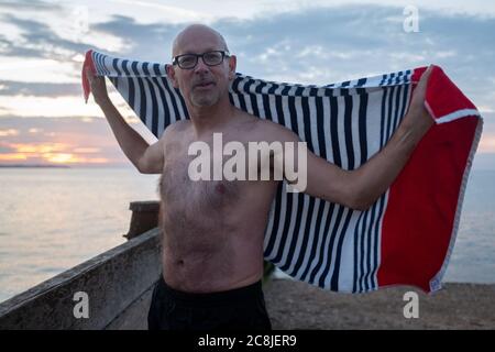 Alors que le soleil se couche sur la lumière du jour et les eaux calmes de l'estuaire de la Tamise, un nageur de mer sauvage se sèche avec une serviette de plage après sa baignade régulière en soirée, le 18 juillet 2020, à Whitstable, Kent, Angleterre. Banque D'Images