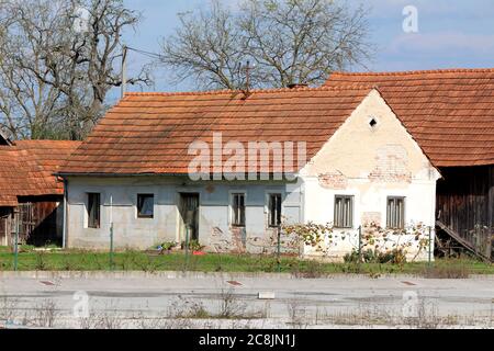 Petite ancienne brique rouge allongée Suburban maison familiale avec craquelé façade et fenêtres en bois délabrées entourées de grands arbres Banque D'Images