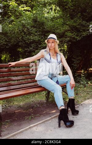 Une fille blonde portant un chapeau, un Jean bleu et des bottes à talons hauts est assise sur un banc dans un parc ou une forêt Banque D'Images