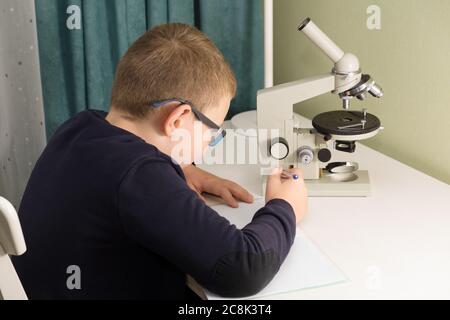 garçon dans des verres et une veste se trouve à une table et fait une tâche d'école Banque D'Images