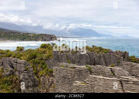 Le célèbre Pancake Rocks sur Dolomite point avec une vue sur la mer vers les montagnes couvertes de nuages. Dolomite point, Île du Sud, Nouvelle-Zélande Banque D'Images