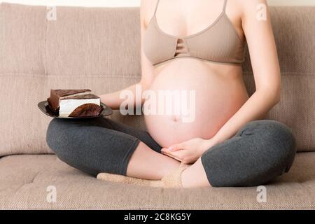 Gros plan de la femme enceinte affamée mange un morceau de gâteau savoureux se détendant sur le canapé à la maison. Envies de douceur pendant la grossesse. Banque D'Images