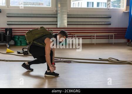 Un instructeur motivé s'agenouille et attache ses chaussures avec un sac à dos et un masque d'entraînement. Commencer la formation. Pour le camp de chaussures et le concept de gym. Banque D'Images