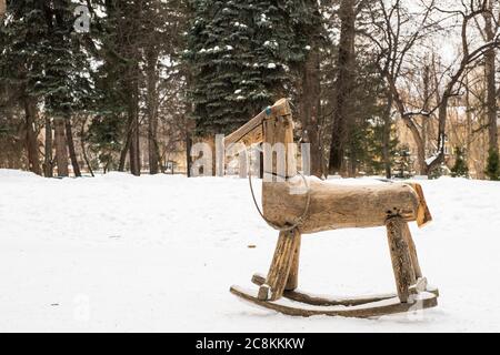 Paysage d'hiver. Un petit balancement en bois pour enfants - un joli cheval dans un clairière sur fond de neige blanche et d'arbres dans un parc de la ville. Banque D'Images