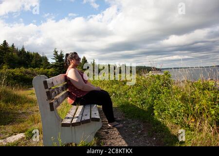 Femme en lunettes de soleil regarde contentement vers l'océan Atlantique tout en étant assise sur un banc de parc Banque D'Images