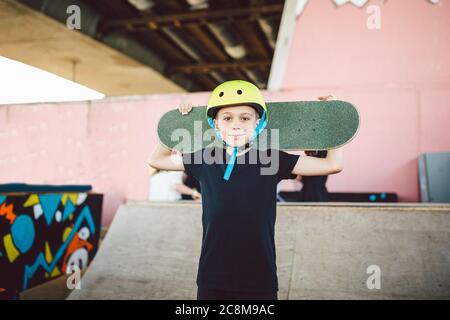 Un skate d'enfant caucasien posant avec un skate board sur les épaules d'un parc de skate extérieur. Portrait de garçon est engagé dans le skateboard sur la rampe à demi-pipe Banque D'Images