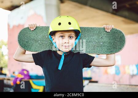 Garçon tenant le skateboard derrière sa tête. Un adorable petit sourire en plein air avec un skateboard. Concept d'enfance, de lixiviation, de style de vie. Enfant urbain avec skate Banque D'Images