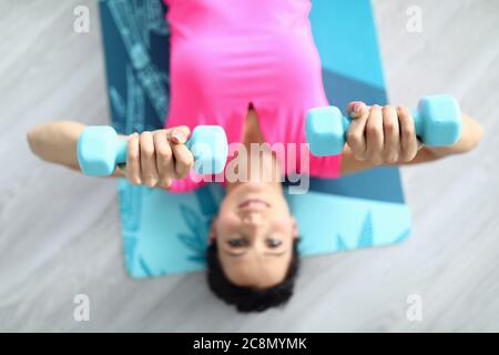 Belle femme en T-shirt rose s'allonger sur le tapis de gymnastique et rais ses bras avec des haltères au-dessus d'elle-même. Banque D'Images