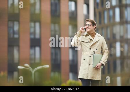 Prenez la taille d'un jeune homme d'affaires souriant parlant avec un smartphone à l'extérieur tout en marchant vers l'appareil photo dans un cadre urbain, dans un espace de copie Banque D'Images