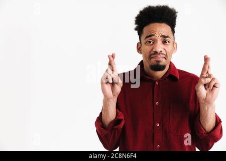 Photo d'un jeune homme africain nerveux posant isolé sur fond de mur blanc montrant les doigts croisés, s'il vous plaît geste Banque D'Images