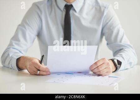 Portrait réduit minimal de jeune homme d'affaires méconnu tenant un document et pendant qu'il travaille assis au bureau et au bureau contre un mur blanc, copie Banque D'Images