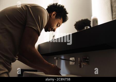 Photo du jeune homme afro-américain avec la barbe en train de nettoyer son visage devant le miroir dans la salle de bains Banque D'Images