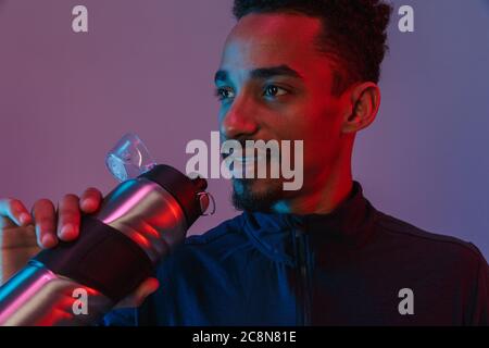 Portrait of smiling african american man in sportswear drinking from thermos isolé sur fond violet Banque D'Images