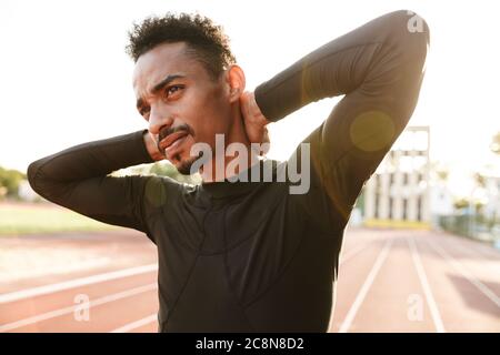 Image of african american man in sportswear touchant son cou en faisant de l'exercice dans stadium de matin à l'extérieur Banque D'Images