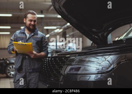 Attention sélective sur une voiture légère, gaie barbu mécanicien écrit sur son bloc-notes sur l'arrière-plan. Un technicien automobile professionnel répare les voitures Banque D'Images