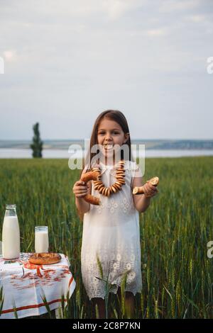 Mignonne souriante petite fille mangeant bagel avec lait dans le champ vert, l'été Banque D'Images